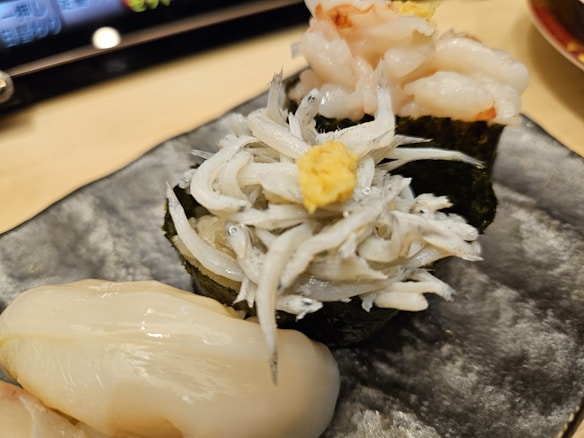 A sushi plate with various types of seafood including delicate slices of shrimp and small white fish, presented on a dark rectangular plate. The sushi is garnished with a small amount of ginger, and a digital screen is blurred in the background.
