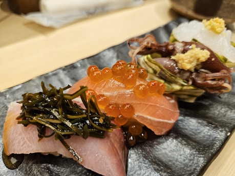 A beautifully presented seafood dish features an assortment of sushi elements. There is a piece of sashimi topped with seaweed, another with bright orange salmon roe, and a third with small squid garnished with ginger on a textured dark plate.
