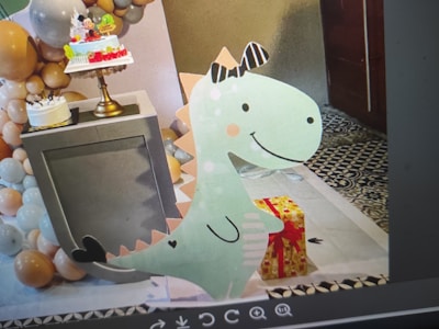 A festive scene with a dinosaur-themed decoration in front of a display table holding two cakes. The background is adorned with a large arrangement of balloons in shades of white, brown, and peach. A colorful gift box with a red ribbon is placed next to the dinosaur cutout, creating a cheerful and playful setting.