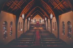 A spacious and empty church interior with high, vaulted wooden ceilings and rows of wooden pews. Stained glass windows line the walls on both sides, allowing colored light to filter through. Elegant, large hanging lanterns illuminate the space, and a pulpit is visible at the far end, positioned behind an altar.