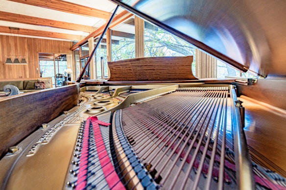 A detailed view of the inside of a grand piano, emphasizing the strings and intricate mechanical components. The piano is situated in a warm, cozy room with wooden walls and large windows that provide a view of greenery outside.