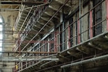 A dilapidated, multi-tiered prison block with metal railings and rusted bars. The structure is worn and old, with peeling paint and signs of decay. Light filters in from a large arched window, adding a touch of natural illumination.