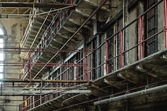 A dilapidated, multi-tiered prison block with metal railings and rusted bars. The structure is worn and old, with peeling paint and signs of decay. Light filters in from a large arched window, adding a touch of natural illumination.