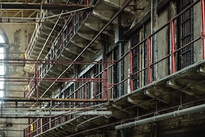 A dilapidated, multi-tiered prison block with metal railings and rusted bars. The structure is worn and old, with peeling paint and signs of decay. Light filters in from a large arched window, adding a touch of natural illumination.