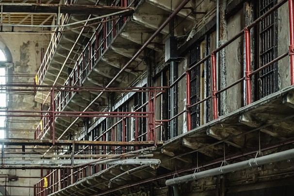 A dilapidated, multi-tiered prison block with metal railings and rusted bars. The structure is worn and old, with peeling paint and signs of decay. Light filters in from a large arched window, adding a touch of natural illumination.