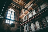 An interior of an old, abandoned prison with multiple levels and barred windows. The building's walls are worn and deteriorating, with visible cracks and peeling paint. Sunlight filters through arched windows, casting shadows across the dimly lit cell blocks. Metal bars and railings line the walkways, accentuated by red piping.