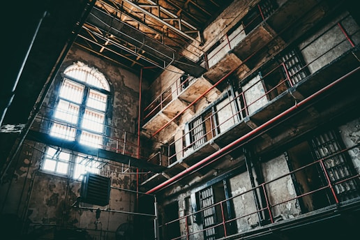 An interior of an old, abandoned prison with multiple levels and barred windows. The building's walls are worn and deteriorating, with visible cracks and peeling paint. Sunlight filters through arched windows, casting shadows across the dimly lit cell blocks. Metal bars and railings line the walkways, accentuated by red piping.
