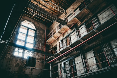 An interior of an old, abandoned prison with multiple levels and barred windows. The building's walls are worn and deteriorating, with visible cracks and peeling paint. Sunlight filters through arched windows, casting shadows across the dimly lit cell blocks. Metal bars and railings line the walkways, accentuated by red piping.