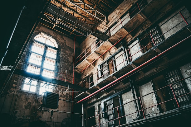 An interior of an old, abandoned prison with multiple levels and barred windows. The building's walls are worn and deteriorating, with visible cracks and peeling paint. Sunlight filters through arched windows, casting shadows across the dimly lit cell blocks. Metal bars and railings line the walkways, accentuated by red piping.