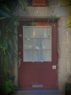 A quaint wooden door with a window is set into a stone wall. The window is divided into small panes and is partially covered by lace curtains. Above the door is a sign that reads 'Portugal.' The doorway is surrounded by lush green plants, creating a cozy and welcoming atmosphere. A number '4' is positioned above the door.