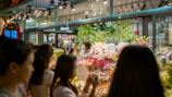 A bustling flower shop with vibrant floral arrangements behind glass. Several people are gathered outside, admiring the array of colorful flowers, including lilies and roses. The shop is well-lit, creating a warm and inviting atmosphere.