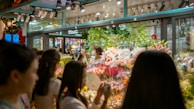 A bustling flower shop with vibrant floral arrangements behind glass. Several people are gathered outside, admiring the array of colorful flowers, including lilies and roses. The shop is well-lit, creating a warm and inviting atmosphere.