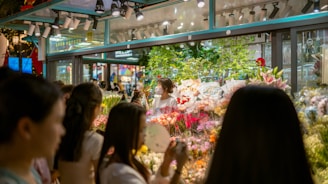 A bustling flower shop with vibrant floral arrangements behind glass. Several people are gathered outside, admiring the array of colorful flowers, including lilies and roses. The shop is well-lit, creating a warm and inviting atmosphere.