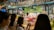 A bustling flower shop with vibrant floral arrangements behind glass. Several people are gathered outside, admiring the array of colorful flowers, including lilies and roses. The shop is well-lit, creating a warm and inviting atmosphere.