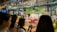 A bustling flower shop with vibrant floral arrangements behind glass. Several people are gathered outside, admiring the array of colorful flowers, including lilies and roses. The shop is well-lit, creating a warm and inviting atmosphere.