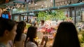 A bustling flower shop with vibrant floral arrangements behind glass. Several people are gathered outside, admiring the array of colorful flowers, including lilies and roses. The shop is well-lit, creating a warm and inviting atmosphere.