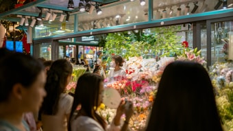 A bustling flower shop with vibrant floral arrangements behind glass. Several people are gathered outside, admiring the array of colorful flowers, including lilies and roses. The shop is well-lit, creating a warm and inviting atmosphere.