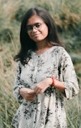 A person with long hair and glasses stands outdoors in a natural setting, wearing a floral-patterned outfit and a red bracelet. The background consists of lush greenery, creating a serene atmosphere.