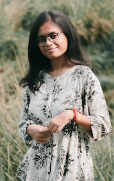 A person with long hair and glasses stands outdoors in a natural setting, wearing a floral-patterned outfit and a red bracelet. The background consists of lush greenery, creating a serene atmosphere.