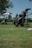 A golf course scene with two golf bags on push carts. One cart has an umbrella attached, and there are some golf balls on the grass. Trees and a clear blue sky are in the background.
