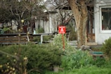 A garden scene with an overcast atmosphere, featuring a bright red warning sign in the center displaying a white skull and crossbones. The sign is surrounded by green shrubbery and appears close to a tree. In the background, there is a wooden bench and a building with visible doors and windows.