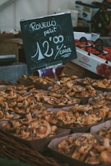 A market display features baskets filled with freshly harvested mushrooms. A chalkboard sign indicates the price and variety of the mushrooms in a casual, handwritten style. Boxes of strawberries are visible in the background, adding to the market ambiance.
