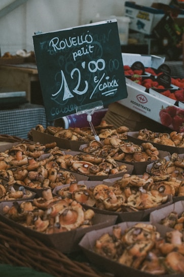 A market display features baskets filled with freshly harvested mushrooms. A chalkboard sign indicates the price and variety of the mushrooms in a casual, handwritten style. Boxes of strawberries are visible in the background, adding to the market ambiance.