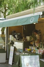 A cozy outdoor cafe with a green awning, featuring a smiling face logo. The counter displays various coffee cups, snacks, and promotional materials. A person stands behind the counter, possibly preparing an order. The setting is shaded by trees, creating a relaxed atmosphere.