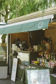 A cozy outdoor cafe with a green awning, featuring a smiling face logo. The counter displays various coffee cups, snacks, and promotional materials. A person stands behind the counter, possibly preparing an order. The setting is shaded by trees, creating a relaxed atmosphere.