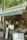 A cozy outdoor cafe with a green awning, featuring a smiling face logo. The counter displays various coffee cups, snacks, and promotional materials. A person stands behind the counter, possibly preparing an order. The setting is shaded by trees, creating a relaxed atmosphere.