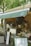 A cozy outdoor cafe with a green awning, featuring a smiling face logo. The counter displays various coffee cups, snacks, and promotional materials. A person stands behind the counter, possibly preparing an order. The setting is shaded by trees, creating a relaxed atmosphere.