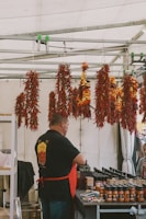 A vendor is standing behind a table at a market stall, with various jars of sauce neatly arranged in front of him. Above, several bunches of drying red and yellow chili peppers are hanging from a metal frame. The vendor is wearing a black shirt with a logo and a red apron. In the background, several customers appear to be looking at the products.