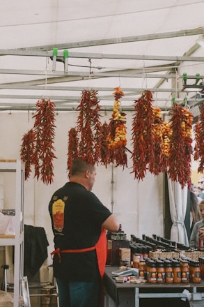 A vendor is standing behind a table at a market stall, with various jars of sauce neatly arranged in front of him. Above, several bunches of drying red and yellow chili peppers are hanging from a metal frame. The vendor is wearing a black shirt with a logo and a red apron. In the background, several customers appear to be looking at the products.