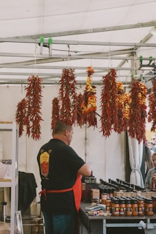 A vendor is standing behind a table at a market stall, with various jars of sauce neatly arranged in front of him. Above, several bunches of drying red and yellow chili peppers are hanging from a metal frame. The vendor is wearing a black shirt with a logo and a red apron. In the background, several customers appear to be looking at the products.