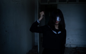 A person with long dark hair, partially covering their face, holds a knife in one hand. They are wearing a dark outfit with a sailor-style collar in a dimly lit, empty room.