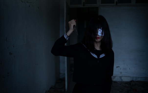 A person with long dark hair, partially covering their face, holds a knife in one hand. They are wearing a dark outfit with a sailor-style collar in a dimly lit, empty room.