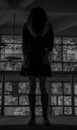 A person stands with their head lowered, obscuring their face, wearing a dark school uniform in front of large windows. The setting is dimly lit, with soft light filtering through the windows.