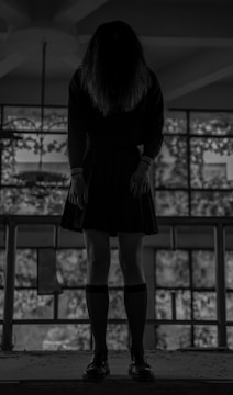 A person stands with their head lowered, obscuring their face, wearing a dark school uniform in front of large windows. The setting is dimly lit, with soft light filtering through the windows.