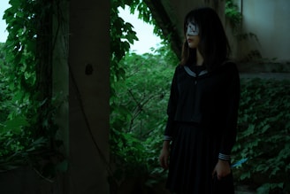 A young woman stands in an overgrown, abandoned area surrounded by lush green foliage. She is wearing a dark school uniform, and her face is partially obscured by a white cloth with red markings. In her right hand, she holds a knife, and her expression conveys a mysterious and eerie presence.