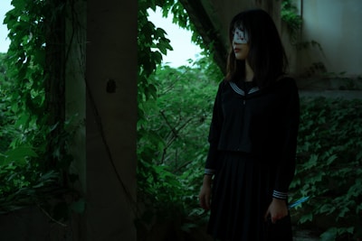 A young woman stands in an overgrown, abandoned area surrounded by lush green foliage. She is wearing a dark school uniform, and her face is partially obscured by a white cloth with red markings. In her right hand, she holds a knife, and her expression conveys a mysterious and eerie presence.
