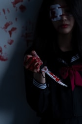 A person wearing a dark outfit with a striped collar holds a blood-stained knife. Their hand is covered in blood, and they have an eye patch over one eye. The background features multiple handprints made with blood.