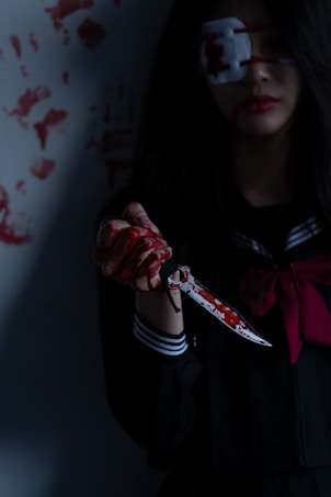 A person wearing a dark outfit with a striped collar holds a blood-stained knife. Their hand is covered in blood, and they have an eye patch over one eye. The background features multiple handprints made with blood.