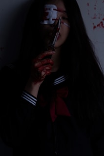 A person with long dark hair holds a bloodied knife close to their face. Their hand and parts of their clothing are stained with blood. The individual wears a dark outfit with a sailor-style collar and a large red bow. An eye patch covers one eye, and there are red splatters on the wall in the background.