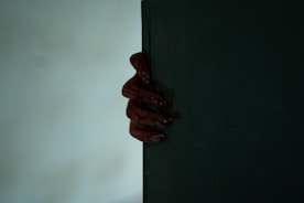 A dark and eerie scene featuring a partially visible hand gripping the edge of a door or wall. The hand is covered in a red substance resembling blood, creating a suspenseful and mysterious atmosphere.