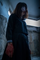 A person stands in a dimly lit, possibly abandoned building with peeling walls. They are wearing a dark outfit and have disheveled hair covering part of their face, which is obscured with a mask or bandage. Their hands are stained with a red substance, possibly representing blood.