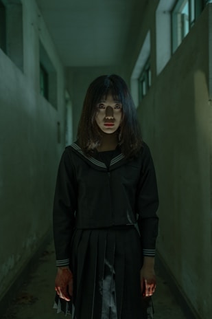 A girl stands in a dimly lit hallway, wearing a dark school uniform with a sailor-style collar. Her face is partially illuminated from below, creating an eerie effect, while her hands appear to be stained, possibly with blood. The atmosphere is tense and unsettling, enhanced by the shadowy corridor with its plain walls and narrow windows allowing minimal light.