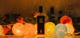 A perfume bottle stands elegantly among a decorative arrangement featuring vibrant roses and spherical string lights. The warm glow of the lights creates a cozy and inviting atmosphere, highlighting the intricate textures of the flowers and the bottle's label.