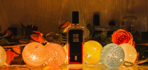 A perfume bottle stands elegantly among a decorative arrangement featuring vibrant roses and spherical string lights. The warm glow of the lights creates a cozy and inviting atmosphere, highlighting the intricate textures of the flowers and the bottle's label.