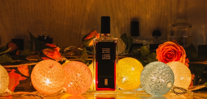 A perfume bottle stands elegantly among a decorative arrangement featuring vibrant roses and spherical string lights. The warm glow of the lights creates a cozy and inviting atmosphere, highlighting the intricate textures of the flowers and the bottle's label.
