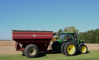 A green tractor is attached to a large red grain cart on a grassy field, with trees and a clear blue sky in the background. The tires on both the tractor and cart are large, suited for agricultural work.